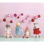 Four plush Christmas-themed dolls are arranged on a mint surface against a soft pink background, with hanging pink and red ornaments above them. The dolls include a nutcracker in pink, a blonde girl in a blue dress, a fairy in pink, and a sheep in a red dress.