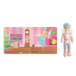 A children's educational book spread shown next to a plush toy nutcracker figure. The book features colorful illustrations of a Christmas tree, a grandfather clock, and a ballerina in a pink room, with text about the Nutcracker.