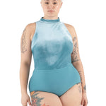 A model with short blonde hair and tattoos wearing a light blue high-neck one-piece leotard, photographed against a solid white background.