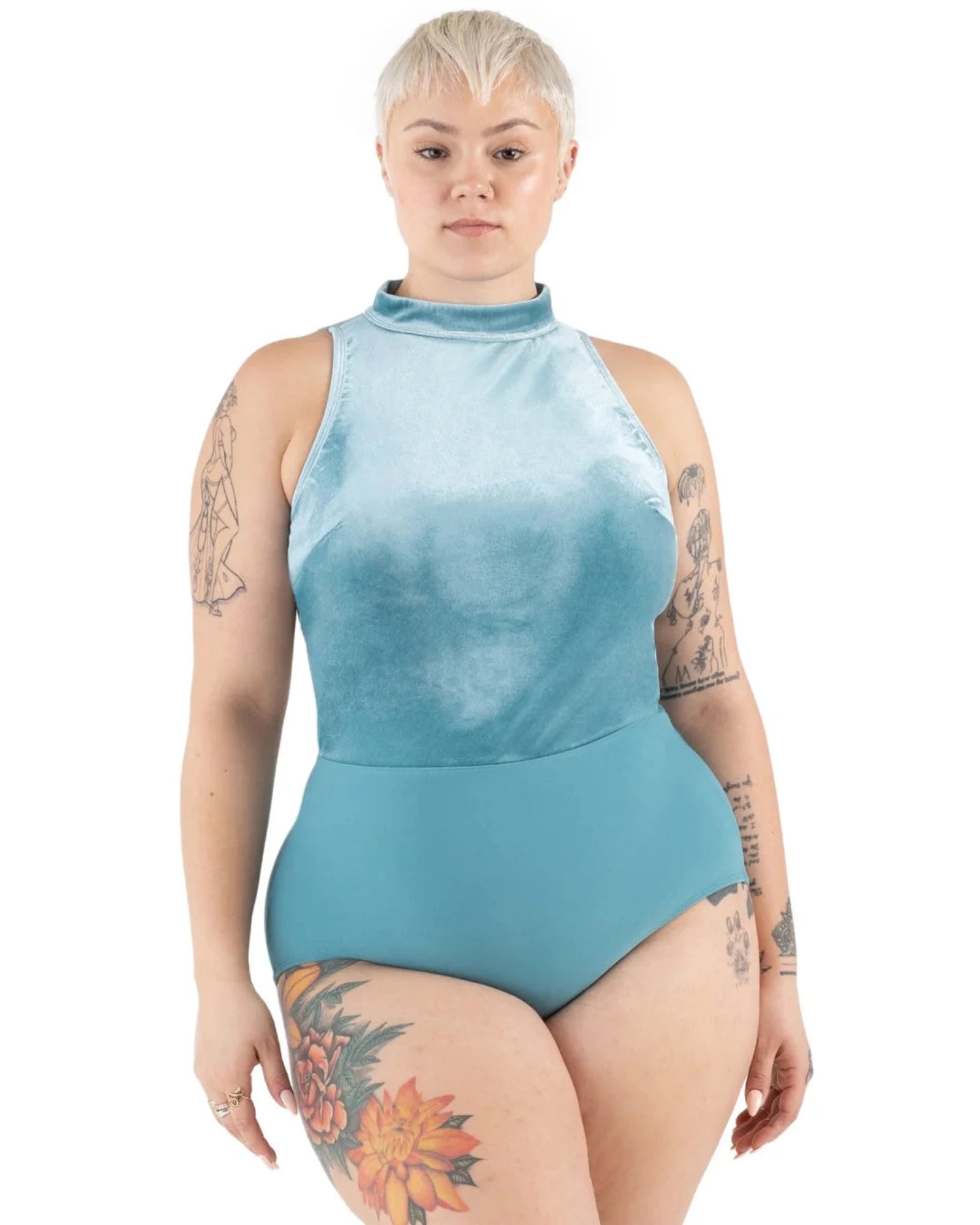 A model with short blonde hair and tattoos wearing a light blue high-neck one-piece leotard, photographed against a solid white background.