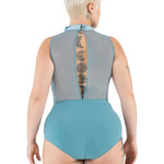 A woman viewed from the back wearing a one-piece leotard with a sheer gray mesh upper back panel and a solid turquoise-blue lower section and leg bands, against a plain white background.