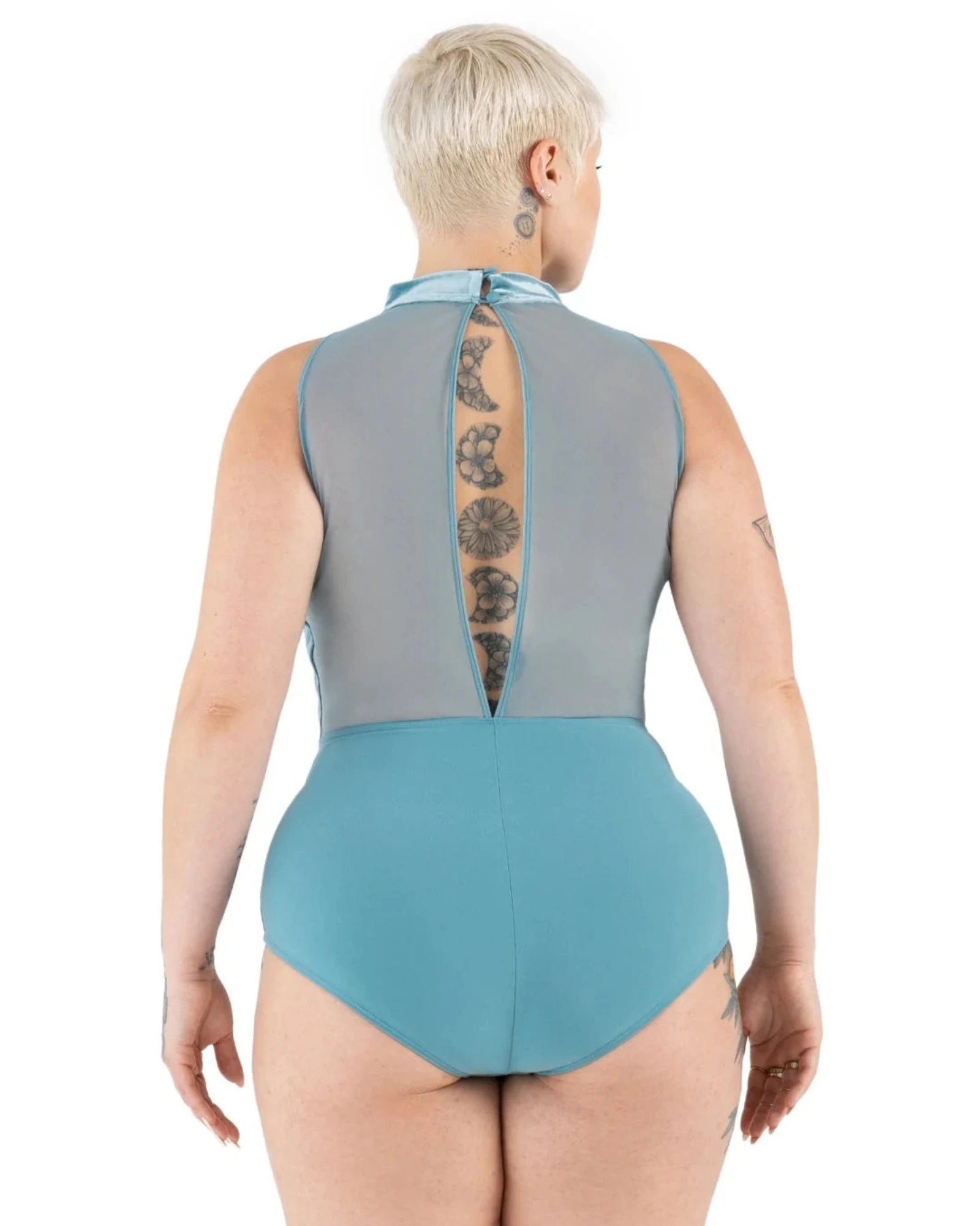 A woman viewed from the back wearing a one-piece leotard with a sheer gray mesh upper back panel and a solid turquoise-blue lower section and leg bands, against a plain white background.