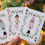 A hand holds three Christmas-themed playing cards featuring Queen, King, and Jack illustrations with green leaf borders and red flowers, set against a blurred Christmas tree background with warm lights.