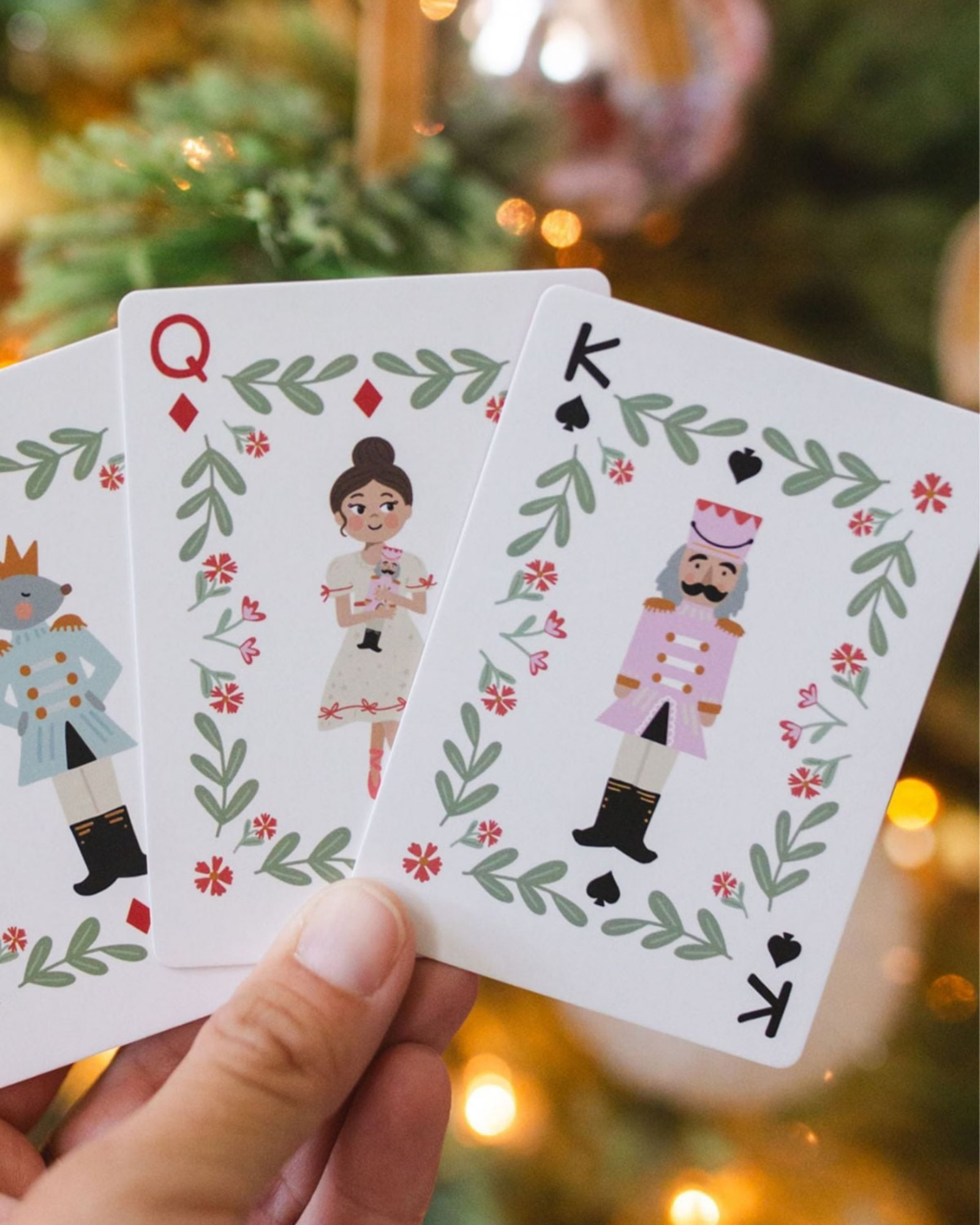 A hand holds three Christmas-themed playing cards featuring Queen, King, and Jack illustrations with green leaf borders and red flowers, set against a blurred Christmas tree background with warm lights.