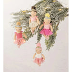 Four ballerina ornaments hanging from a decorated evergreen branch against a plain off-white wall. The ornaments feature soft pink and white tutus, delicate bead hair, and small warm fairy lights.