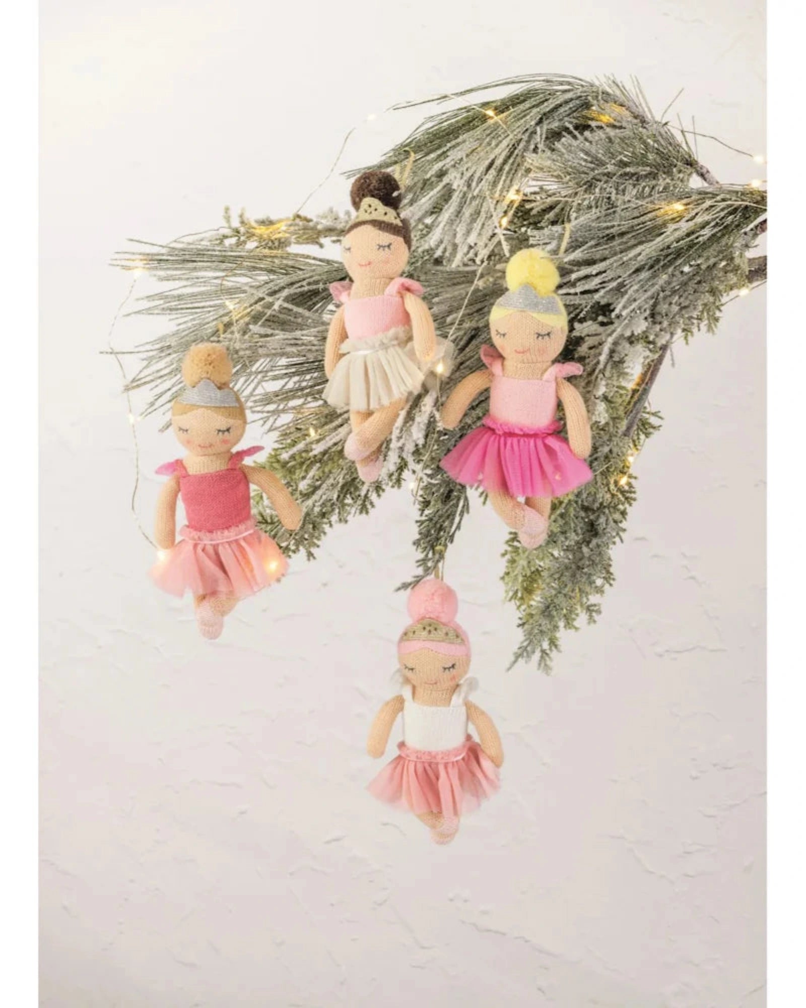 Four ballerina ornaments hanging from a decorated evergreen branch against a plain off-white wall. The ornaments feature soft pink and white tutus, delicate bead hair, and small warm fairy lights.