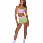 A young girl modeling a two-piece activewear set consisting of a pink and light green polka dot crop top and matching light green shorts, paired with light purple socks, against a solid white background.