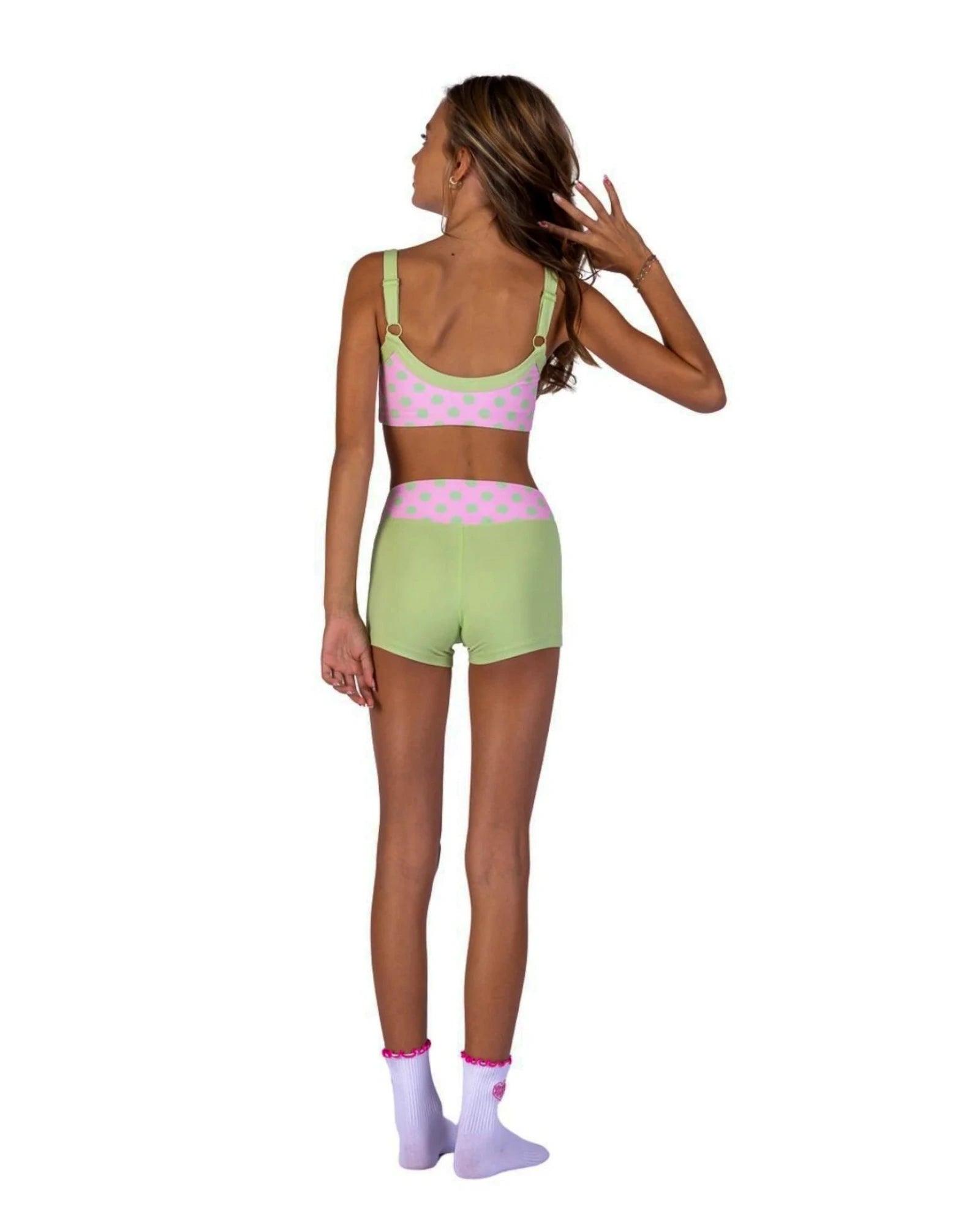 A woman shown from the back wearing a pastel two-piece dance set with a pink and light green polka dot bra topand matching shorts, paired with light purple socks, on a solid white background.