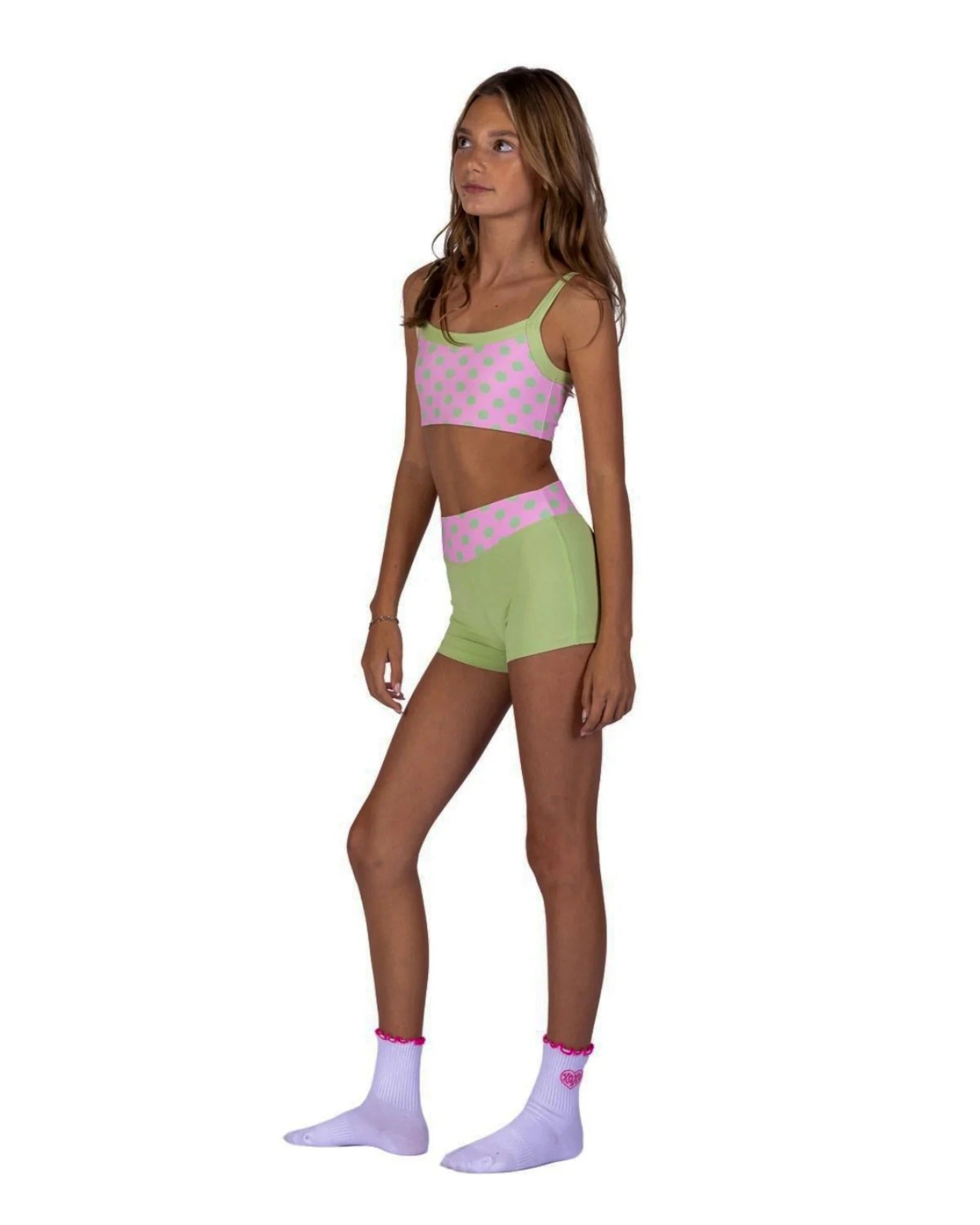 A young girl modeling a matching two-piece activewear set consisting of a pink and green polka dot sports bra and shorts, paired with light purple socks, against a solid white background.