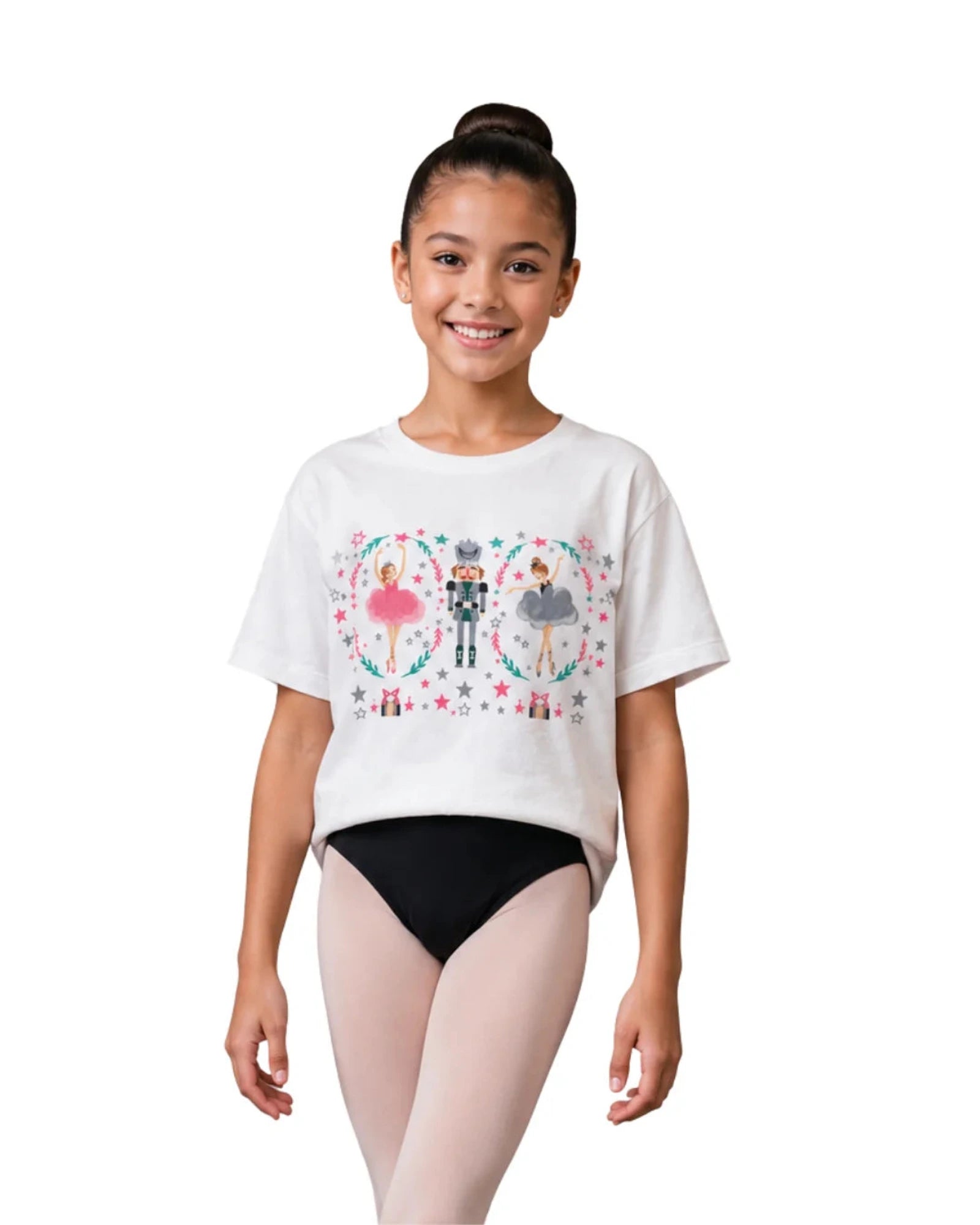 A young girl with her hair in a bun stands against a white background wearing a white graphic t-shirt with a Nutcracker theme and black leotard bottoms.