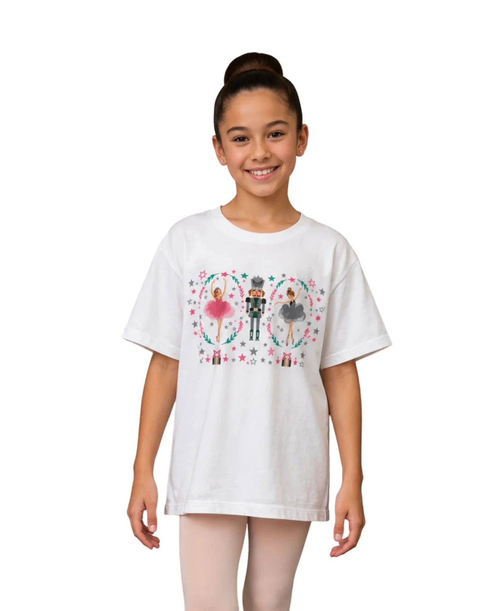 A young girl with her hair in a bun stands against a white background wearing a white oversized graphic t-shirt. The t-shirt features a colorful illustration of a Nutcracker surrounded by hearts and stars.