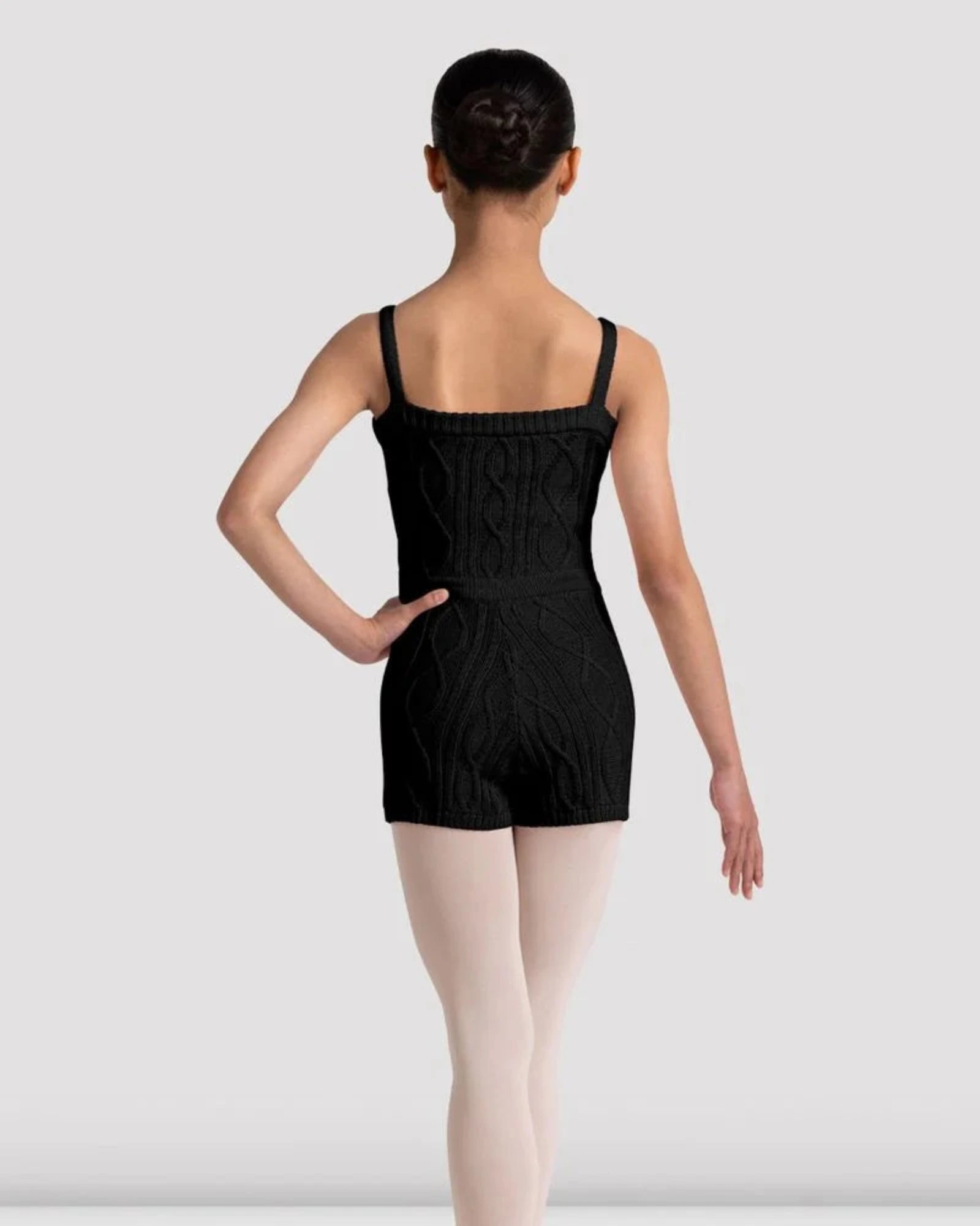 A child dancer shown from the back wearing a black cable-knit unitard, light pink tights, and ballet shoes against a light gray studio background.