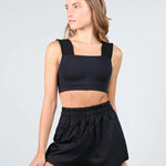A woman modeling a black two-piece dancewear set consisting of a square-neck crop top with thick shoulder straps and matching high-waisted elastic-waist shorts, against a light gray studio background.