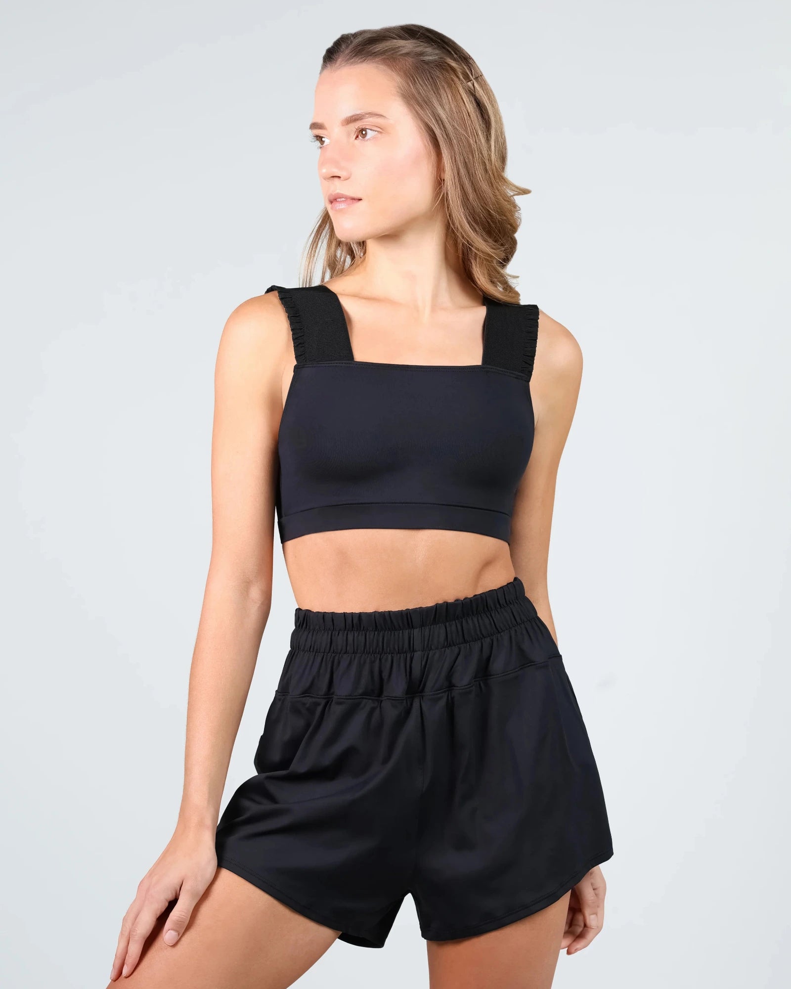 A woman modeling a black two-piece dancewear set consisting of a square-neck crop top with thick shoulder straps and matching high-waisted elastic-waist shorts, against a light gray studio background.