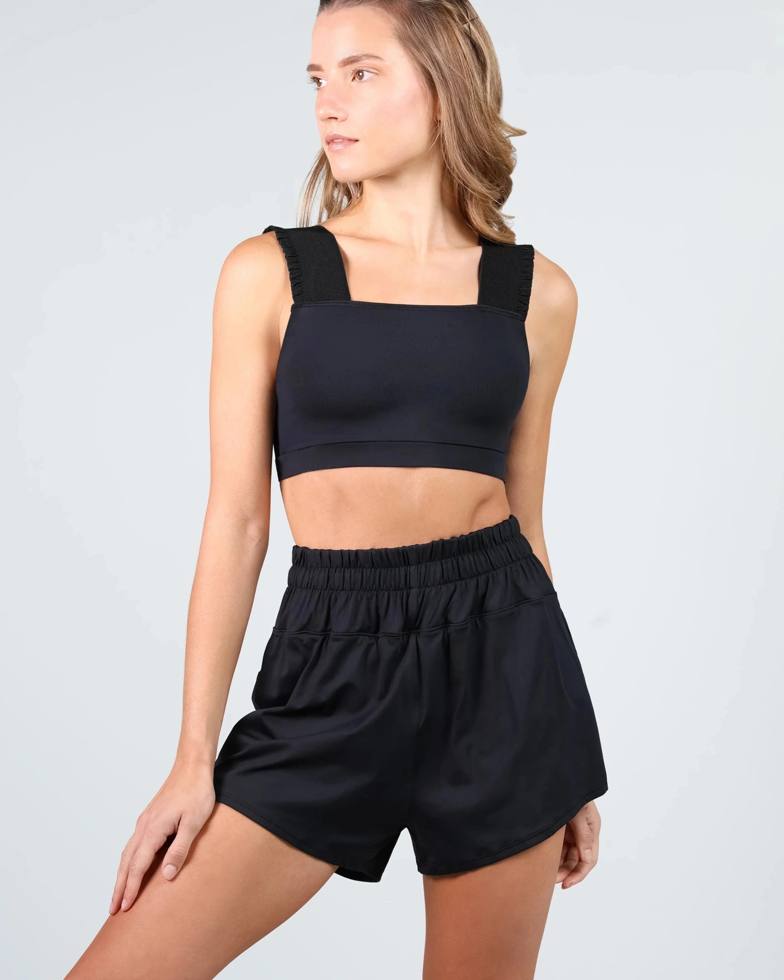 A woman modeling a black two-piece dance set consisting of a square-neck crop top with thick shoulder straps and matching high-waisted elastic-waist shorts, against a light gray studio background.