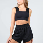 A woman modeling a black two-piece dancewear set consisting of a square-neck crop top with thick shoulder straps and matching high-waisted elastic-waist shorts, against a light gray studio background.