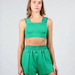 A woman modeling a bright green two-piece dancewear set consisting of a cropped tank top with wide shoulder straps and matching high-waisted shorts with an elastic waistband, shot against a light gray studio background.