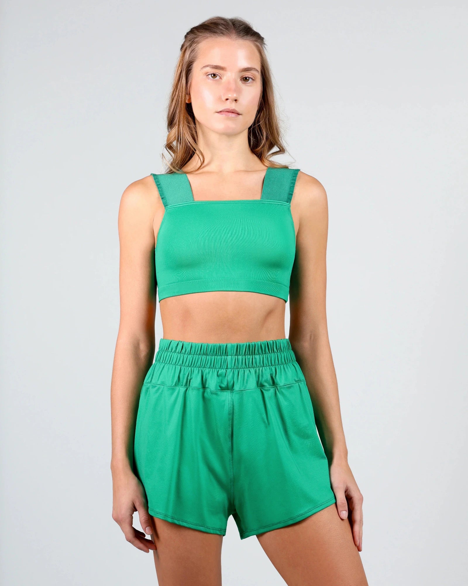 A woman modeling a bright green two-piece dancewear set consisting of a cropped tank top with wide shoulder straps and matching high-waisted shorts with an elastic waistband, shot against a light gray studio background.