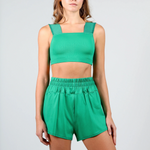A woman modeling a bright green two-piece dance wear set consisting of a cropped tank top with wide shoulder straps and matching high-waisted shorts with an elastic waistband, shot against a light gray studio background.