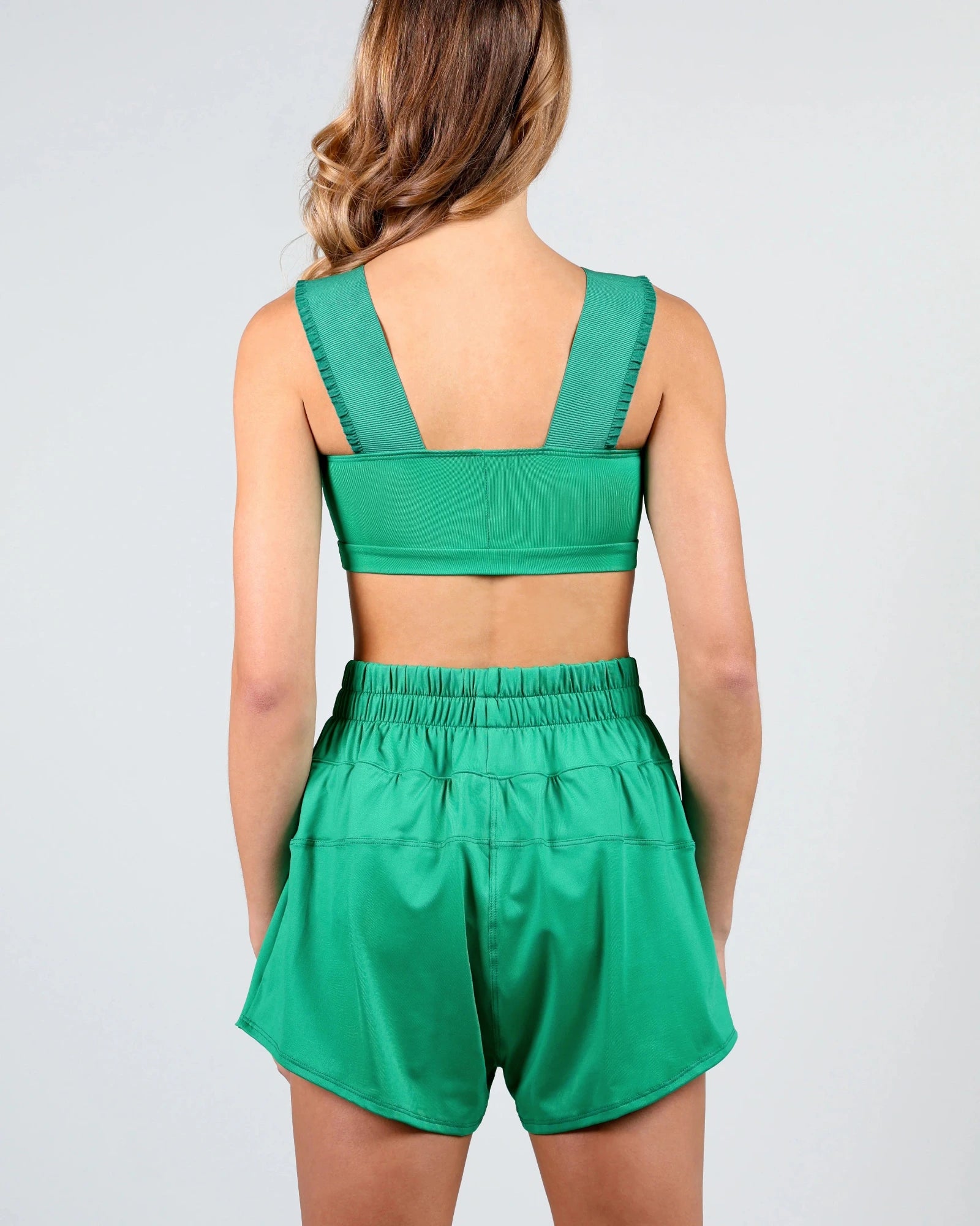 A woman shown from the back wearing a matching green dance set consisting of a cropped tank top with wide shoulder straps and high-waisted shorts with an elastic waistband, against a light gray studio background.