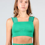 A woman modeling a bright green dance crop top with a square neckline and wide shoulder straps, paired with matching green athletic pants, against a light gray studio background.