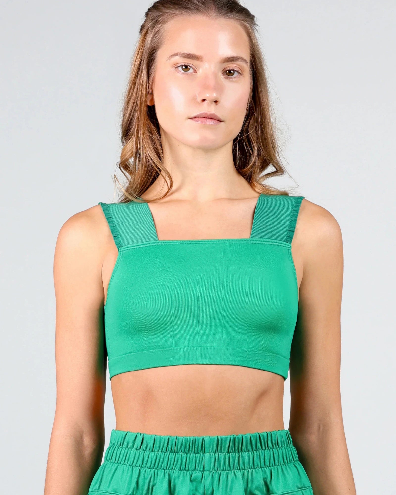 A woman modeling a bright green dance crop top with a square neckline and wide shoulder straps, paired with matching green athletic pants, against a light gray studio background.