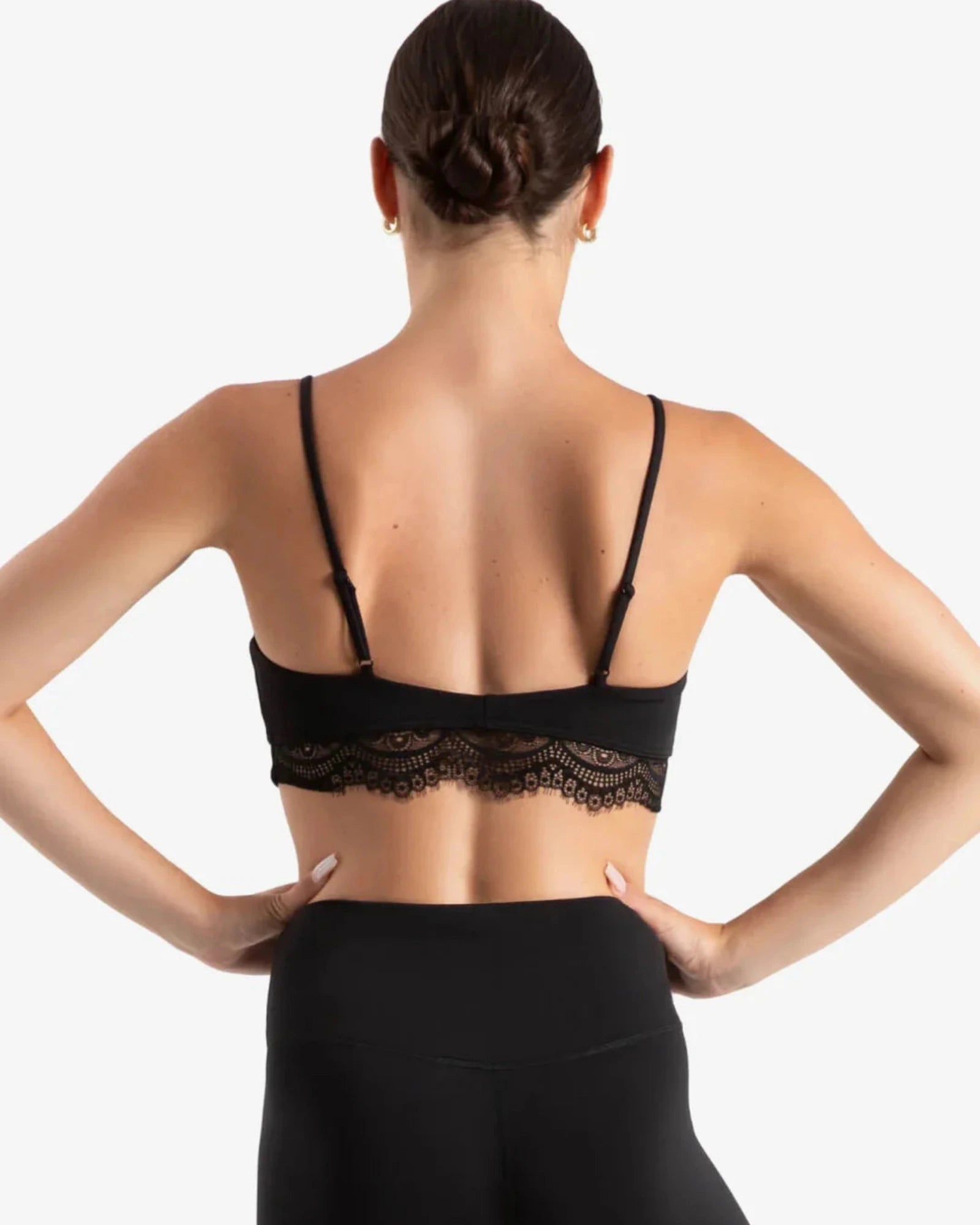 Rear view of a woman wearing a black lace-trimmed dance bra and black leggings against a light gray studio background.