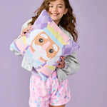 A young girl with long brown hair stands against a solid lavender background, holding a plush pillow with a cartoon prince design. She is wearing a light gray jacket and pink patterned shorts.
