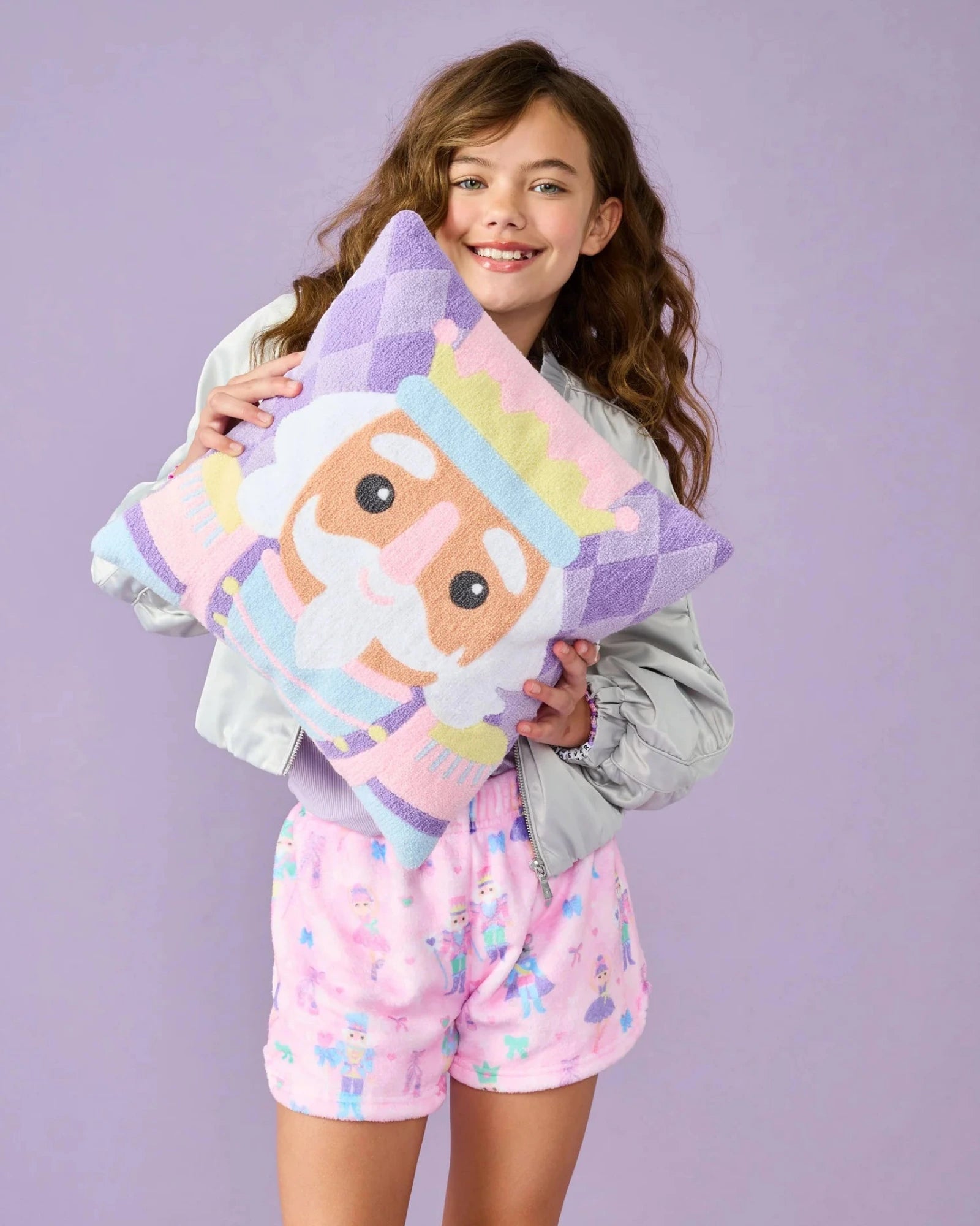 A young girl with long brown hair stands against a solid lavender background, holding a plush pillow with a cartoon prince design. She is wearing a light gray jacket and pink patterned shorts.