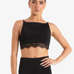 A woman modeling a black dance crop top with a wide neckline and delicate lace trim, paired with black leggings, against a plain light gray studio background.