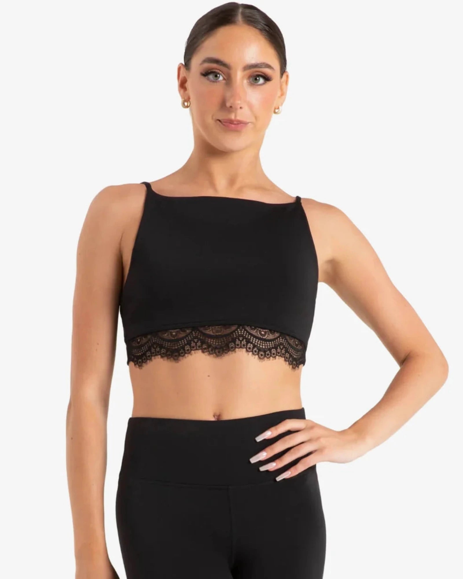 A woman modeling a black dance crop top with a wide neckline and delicate lace trim, paired with black leggings, against a plain light gray studio background.