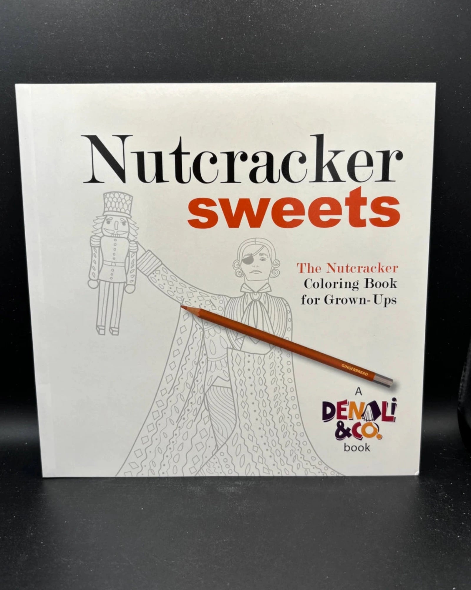 The Nutcracker Sweets coloring book for grown-ups by Denali & Co., featuring a white cover with black and red text and line art of a nutcracker and a child, with a wooden pencil resting across the front, set against a dark background.