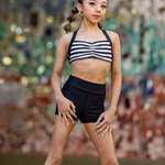 A young girl dancer posing in a black and white striped halter crop top and black high-waisted dance shorts against a colorful, blurred mosaic background.