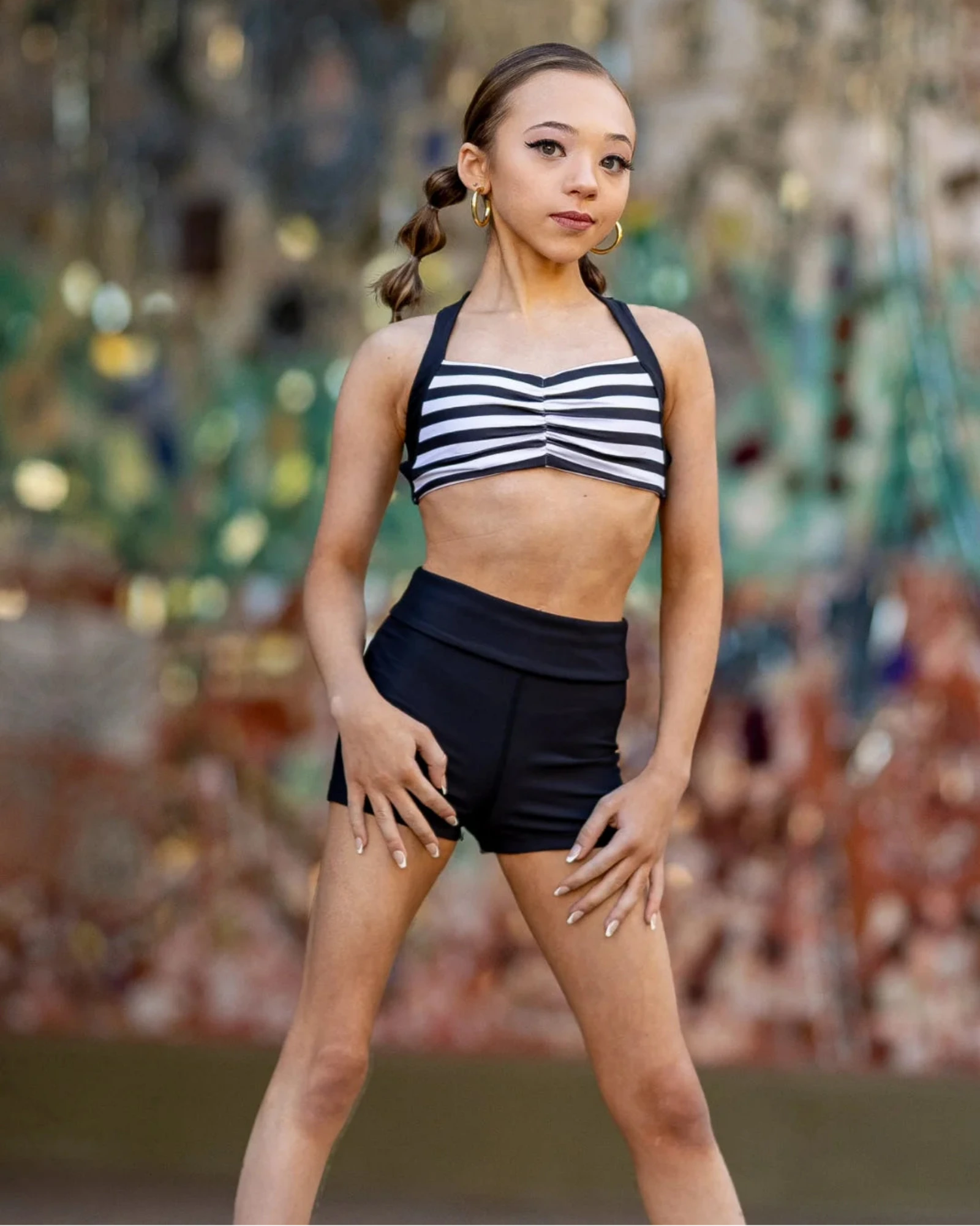 A young girl dancer posing in a black and white striped halter crop top and black high-waisted dance shorts against a colorful, blurred mosaic background.
