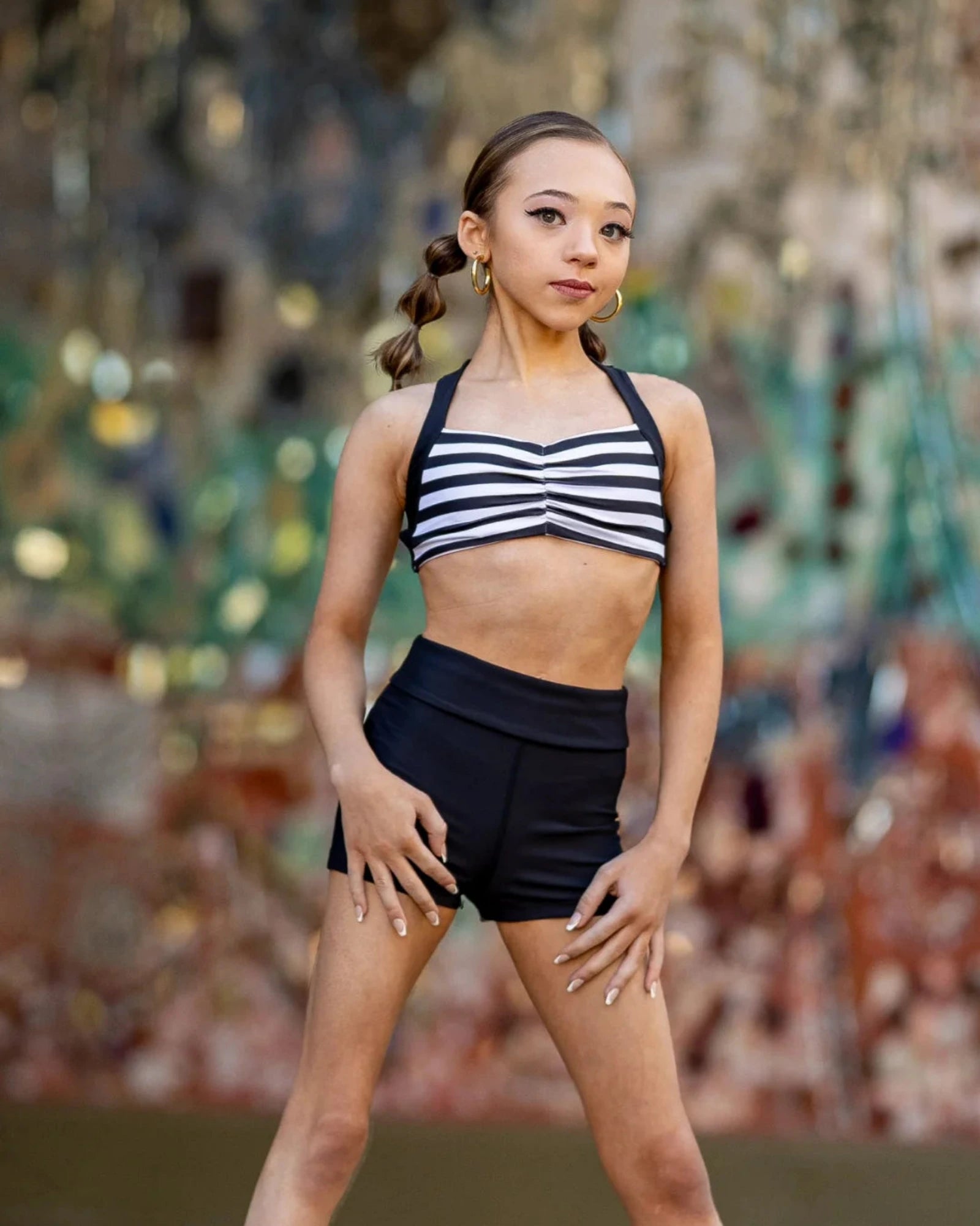 A young girl dancer posing in a black and white striped halter crop top and black high-waisted dance shorts against a colorful, blurred mosaic background.