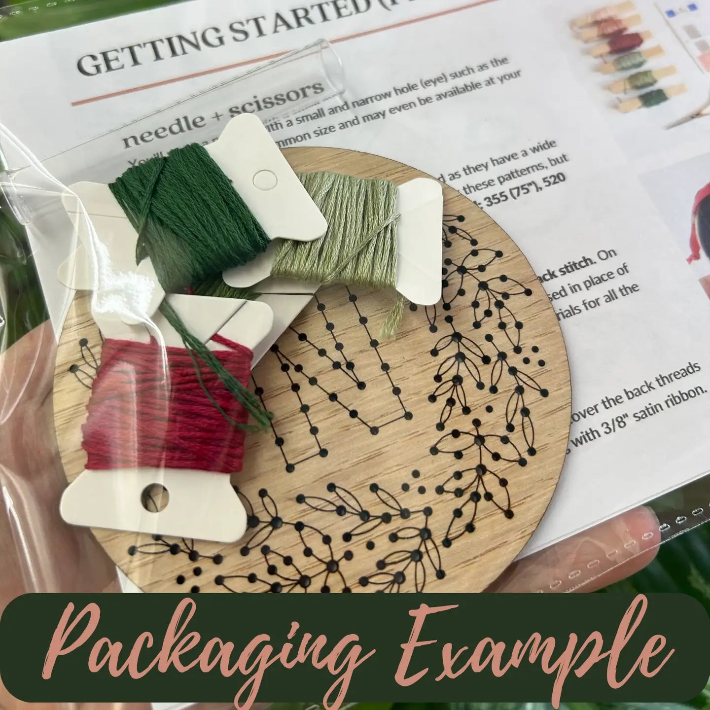 A close-up of a packaged embroidery kit showing a wooden hoop with black stitched leaf outlines, several thread spools in green, red, and beige, and printed instruction sheets behind them. A dark green banner at the bottom reads 'Packaging Example'.