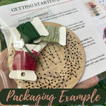 A close-up of a packaged embroidery kit showing a wooden hoop with black stitched leaf outlines, several thread spools in green, red, and beige, and printed instruction sheets behind them. A dark green banner at the bottom reads 'Packaging Example'.