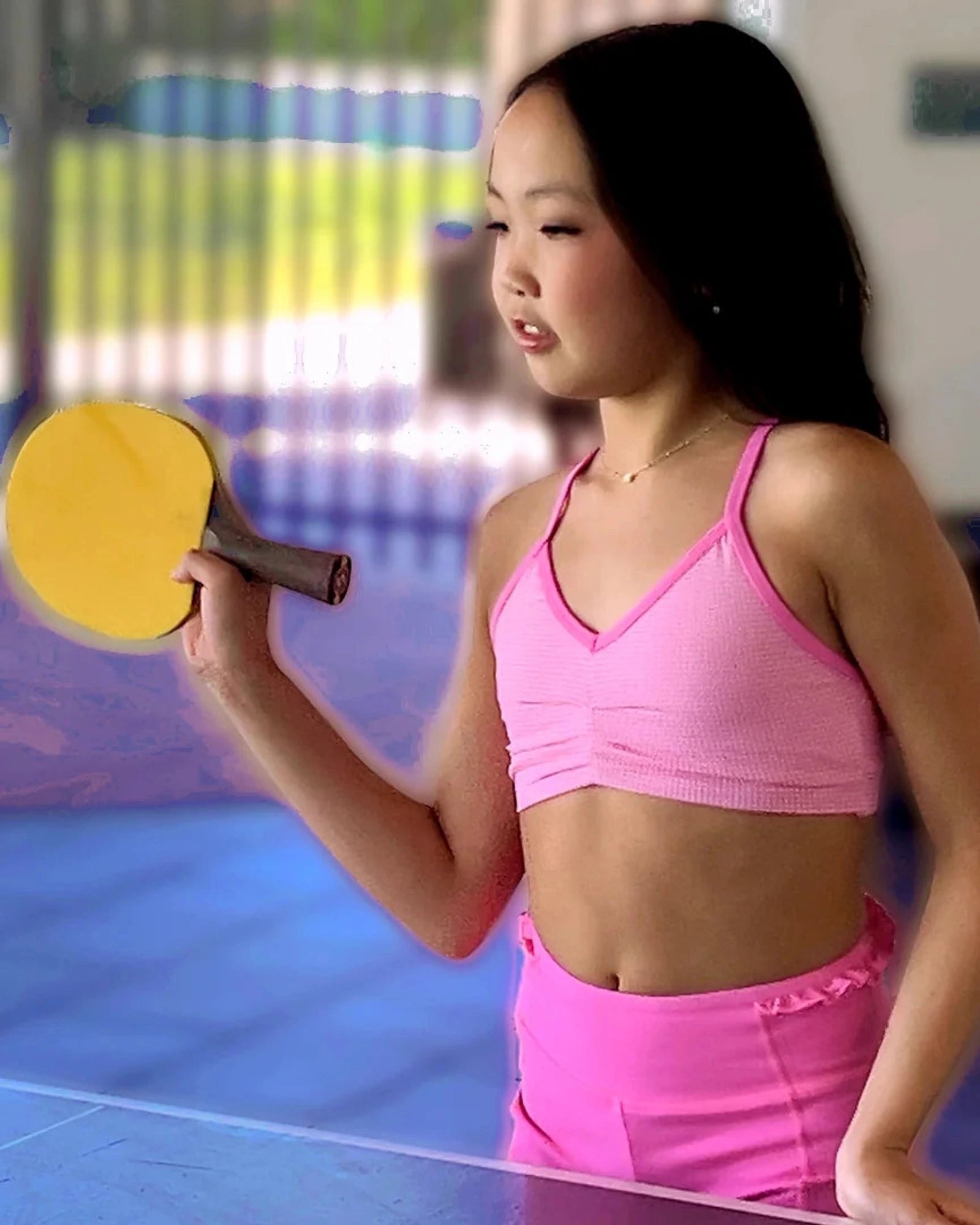 A young girl wearing a pink bra top and matching pink athletic shorts stands on a table tennis table, holding a yellow table tennis paddle.