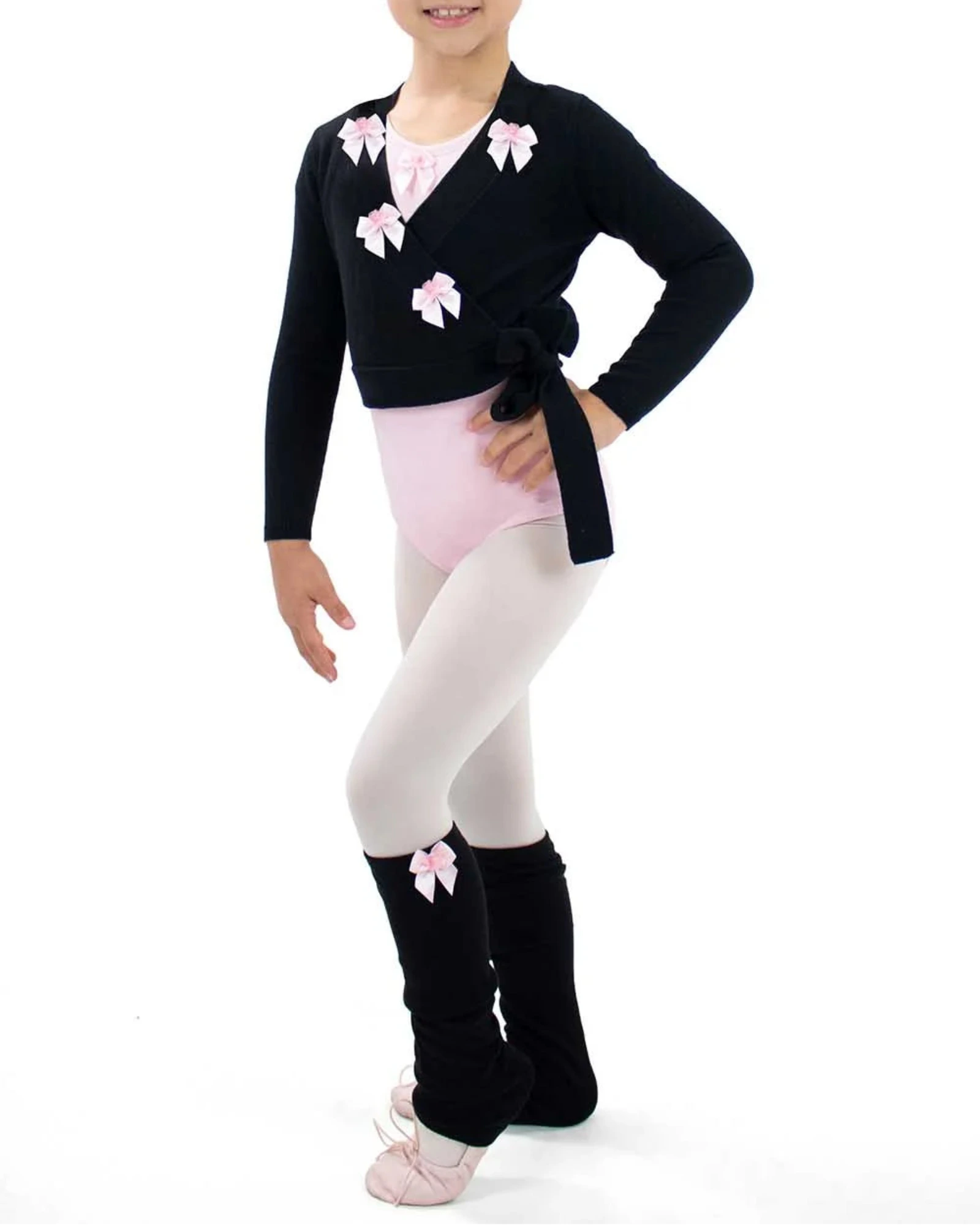 A young girl dancer posing in a black long-sleeve dance cardigan with pink bow details, a light pink leotard, white tights, and black leg warmers with pink bows, set against a plain white background.