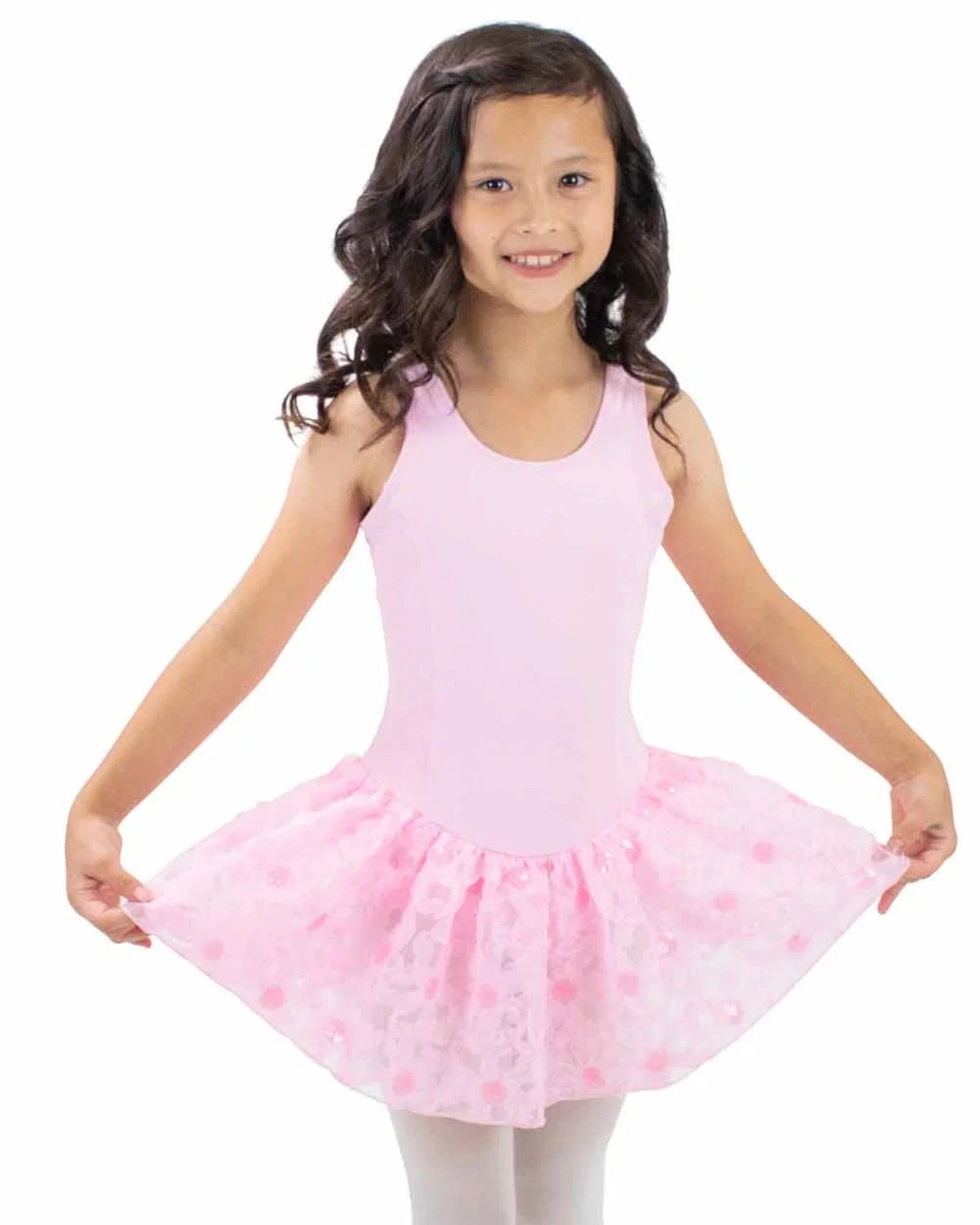 A young girl with long dark curly hair wearing a light pink sleeveless leotard and a matching pink tutu skirt with darker pink flowers, posing against a solid white background.