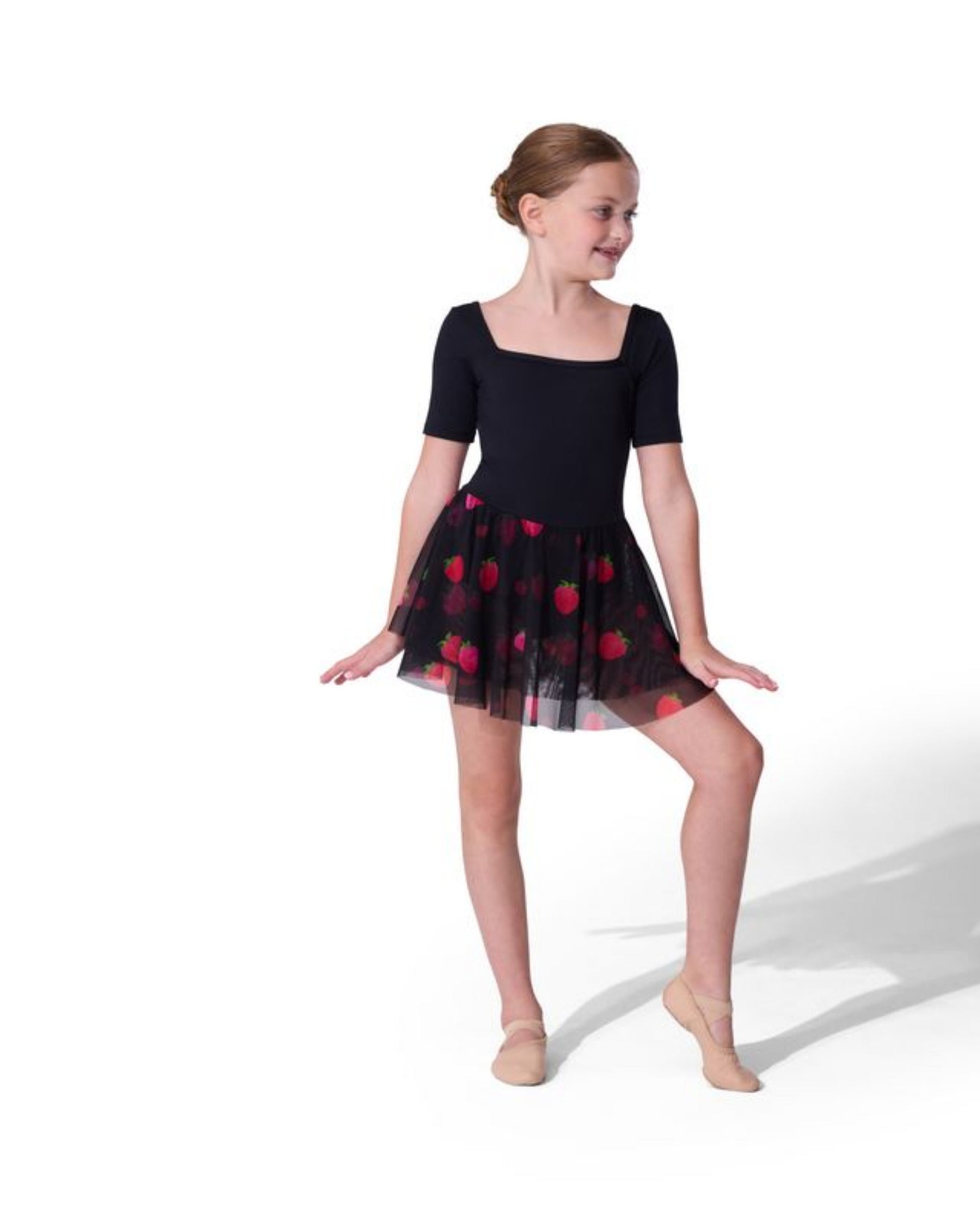 A young girl dancer wearing a black short-sleeve leotard and a black tulle tutu skirt with red strawberries, posed against a solid white studio background.