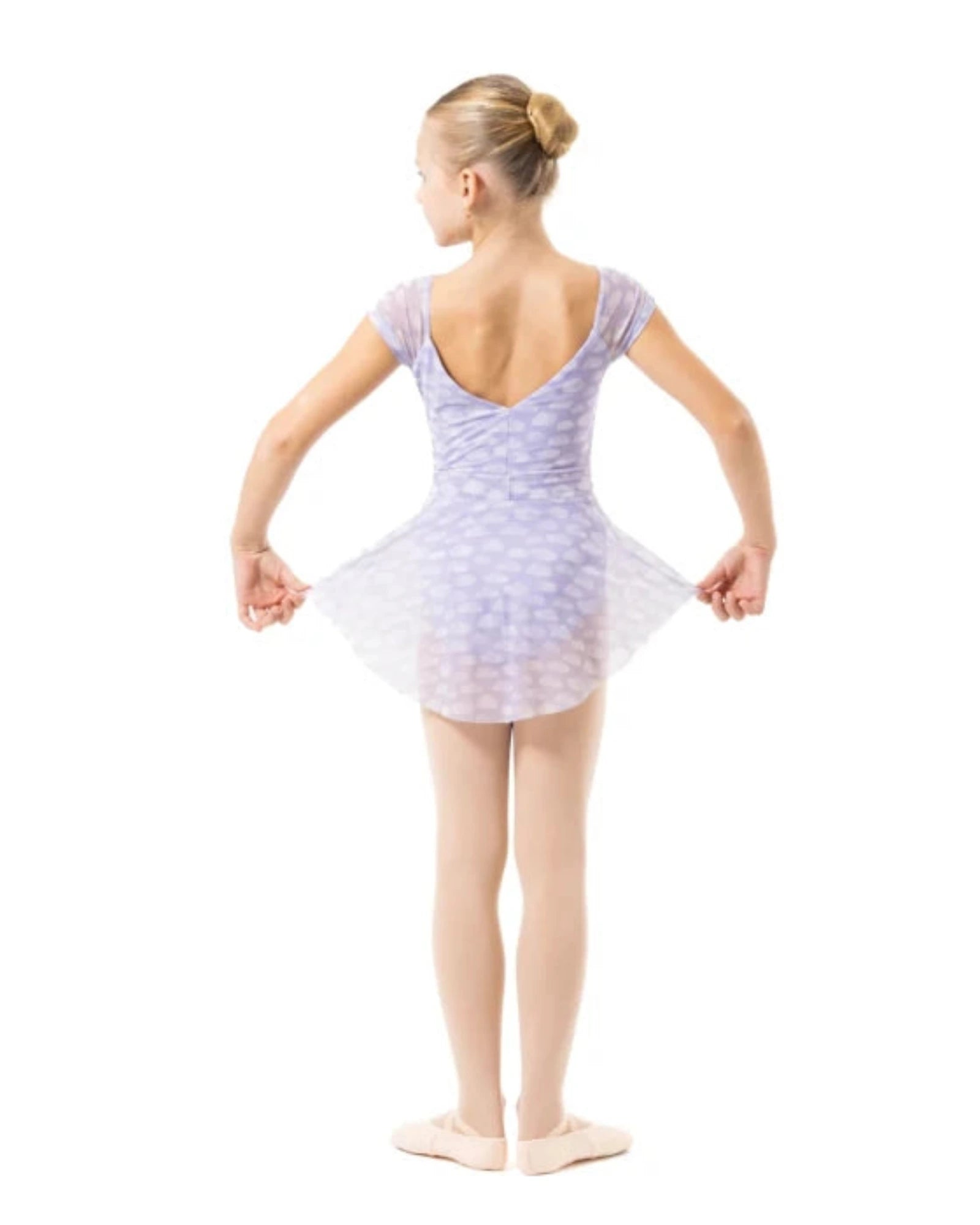 A young girl dancer viewed from behind, wearing a light lavender and white patterned tutu leotard, standing on a solid white background.