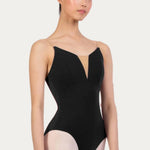 A female dancer wearing a black bodysuit with thin spaghetti straps and a deep V-neckline, paired with light pink tights, set against a plain light gray studio background.