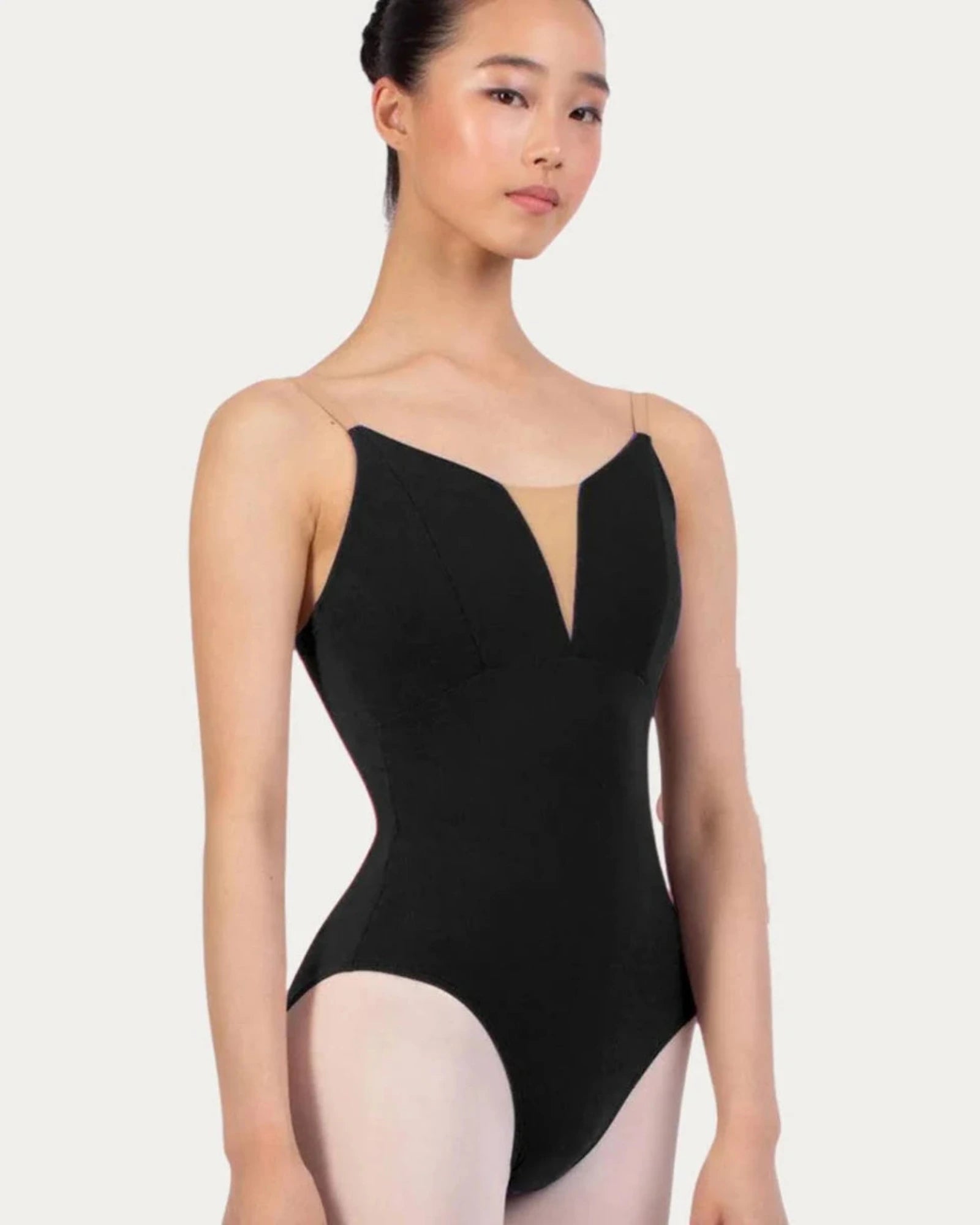 A female dancer wearing a black bodysuit with thin spaghetti straps and a deep V-neckline, paired with light pink tights, set against a plain light gray studio background.