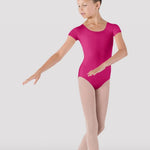 A young girl dancer posing in a bright magenta short-sleeve leotard, light pink tights, and pink pointe shoes against a light gray studio background.