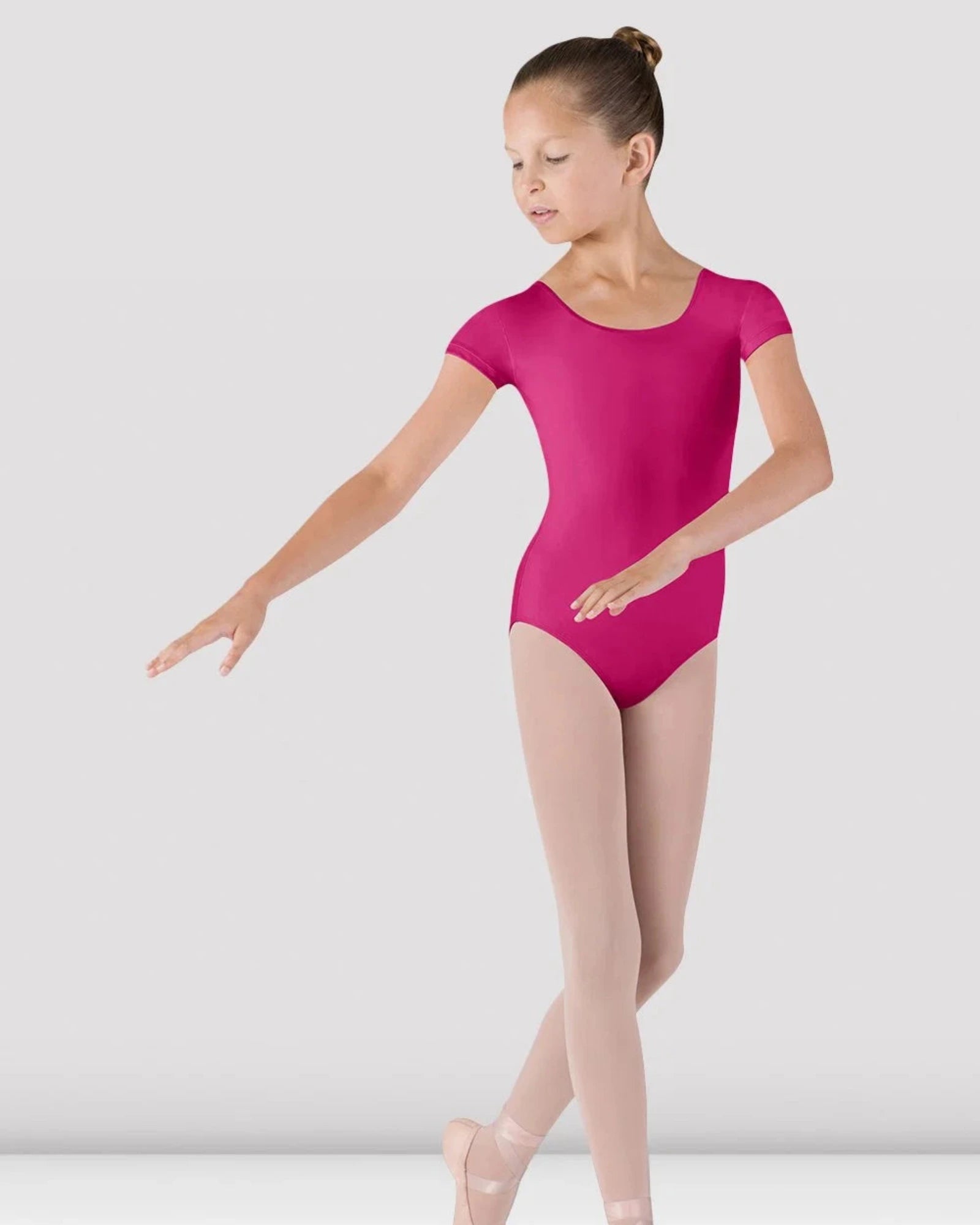 A young girl dancer posing in a bright magenta short-sleeve leotard, light pink tights, and pink pointe shoes against a light gray studio background.