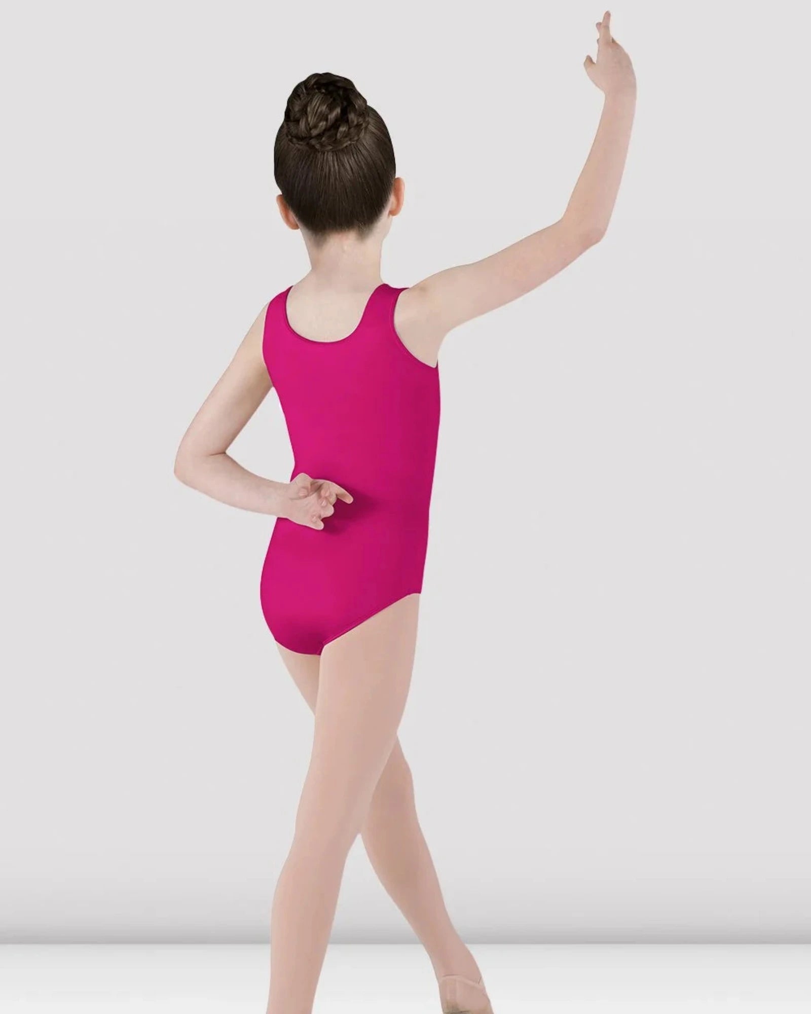 A young girl dancer viewed from behind wearing a bright magenta pink sleeveless leotard and nude tights, posing in a ballet position against a light gray studio background.