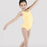 A young girl dancer posing in a tank pale yellow leotard and nude tights against a light gray studio background.