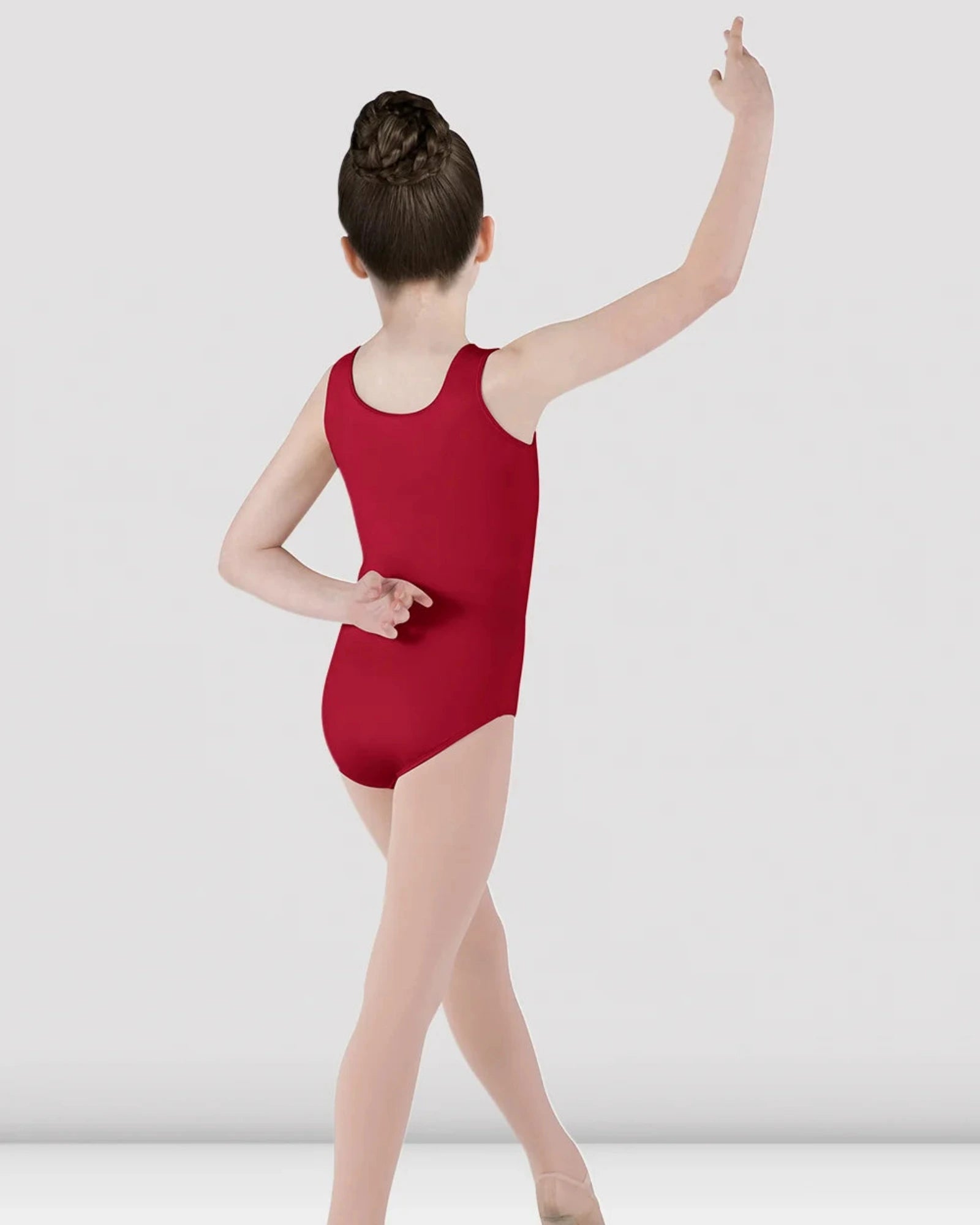 A young girl dancer viewed from behind, wearing a sleeveless deep red leotard and light pink tights, posing in a studio with a light gray background.