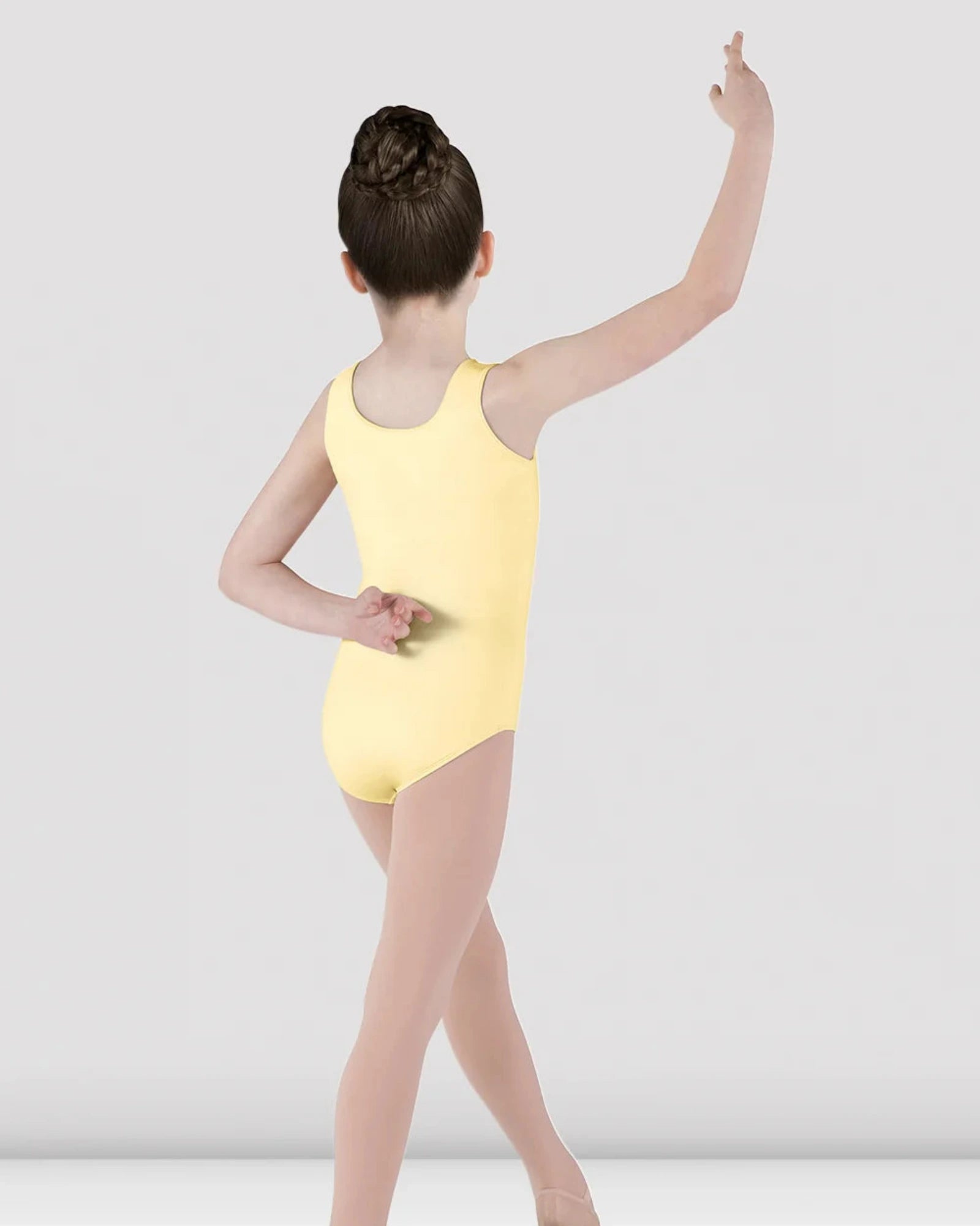 A young girl dancer shown from the back wearing a pale yellow sleeveless leotard and nude tights, posing in a studio setting against a light gray background.